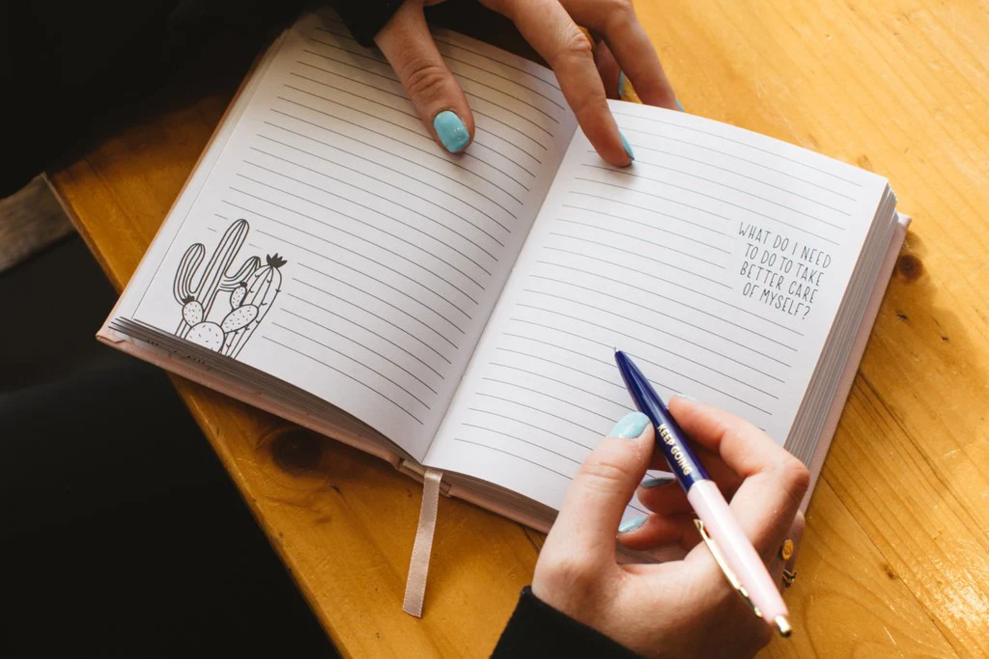 An open book with lined pages is open on a desk. A hand holding a pen sits above the page about to write. Text on the page says “What do I need to do to take better care of myself?”