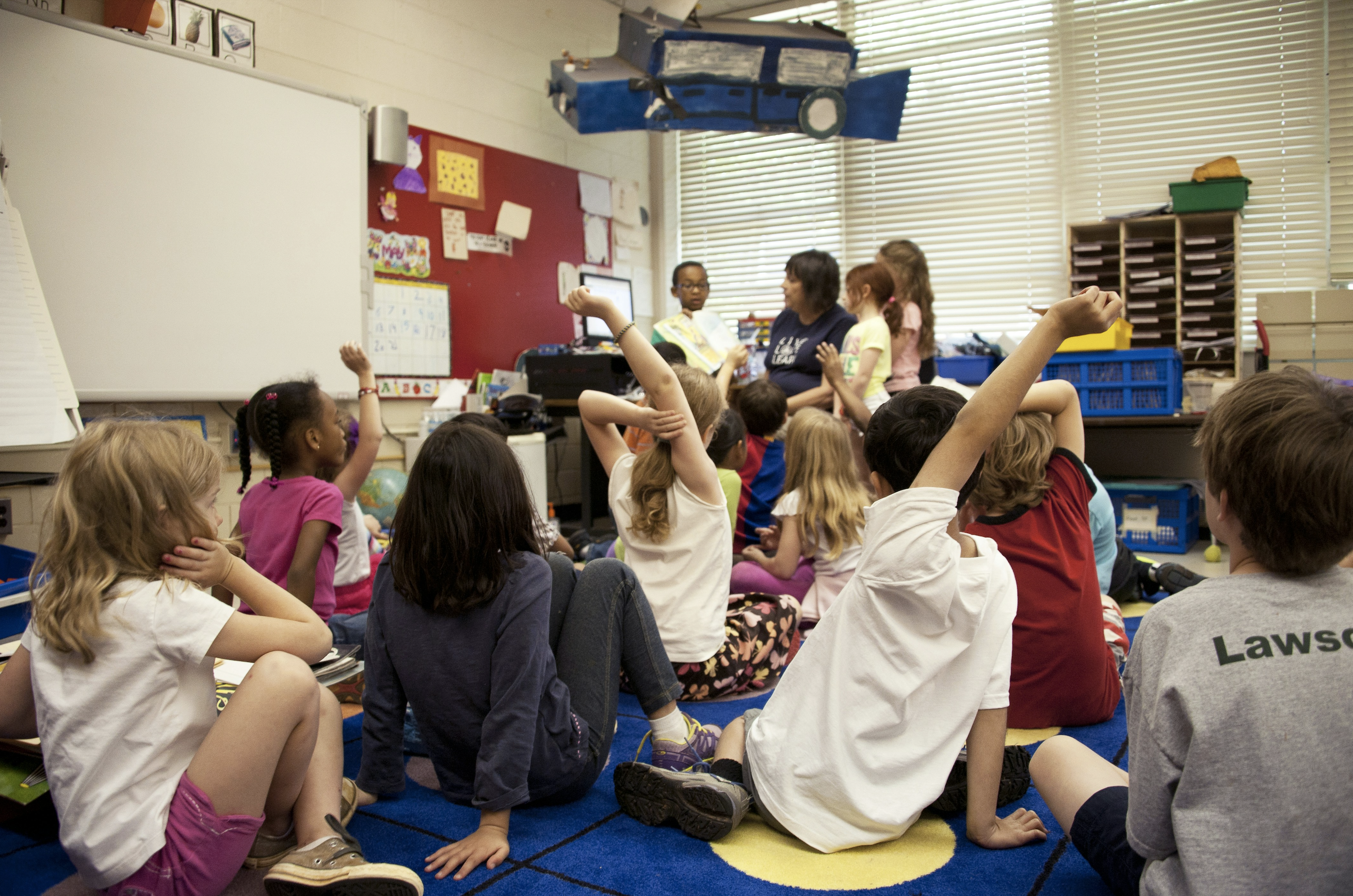 Children in classroom sit on carpet listening to their teacher